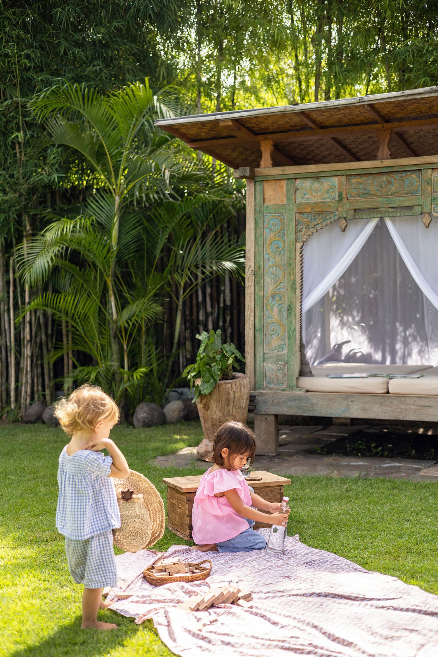 Duas crianças brincando em um cobertor em um jardim com uma estrutura de madeira e uma grande planta em vaso.