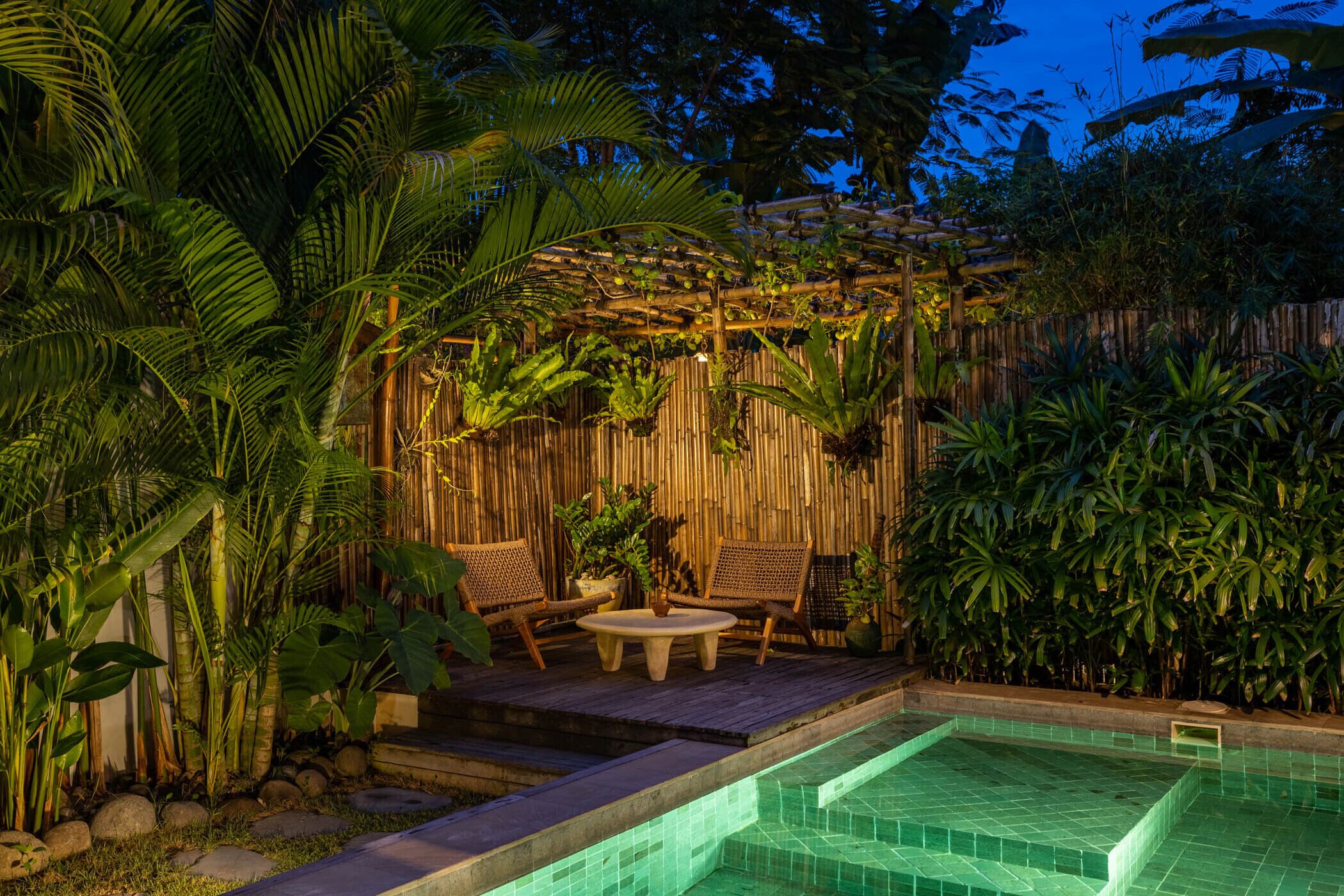 A pool area with a wooden deck, seating, and lush tropical plants.