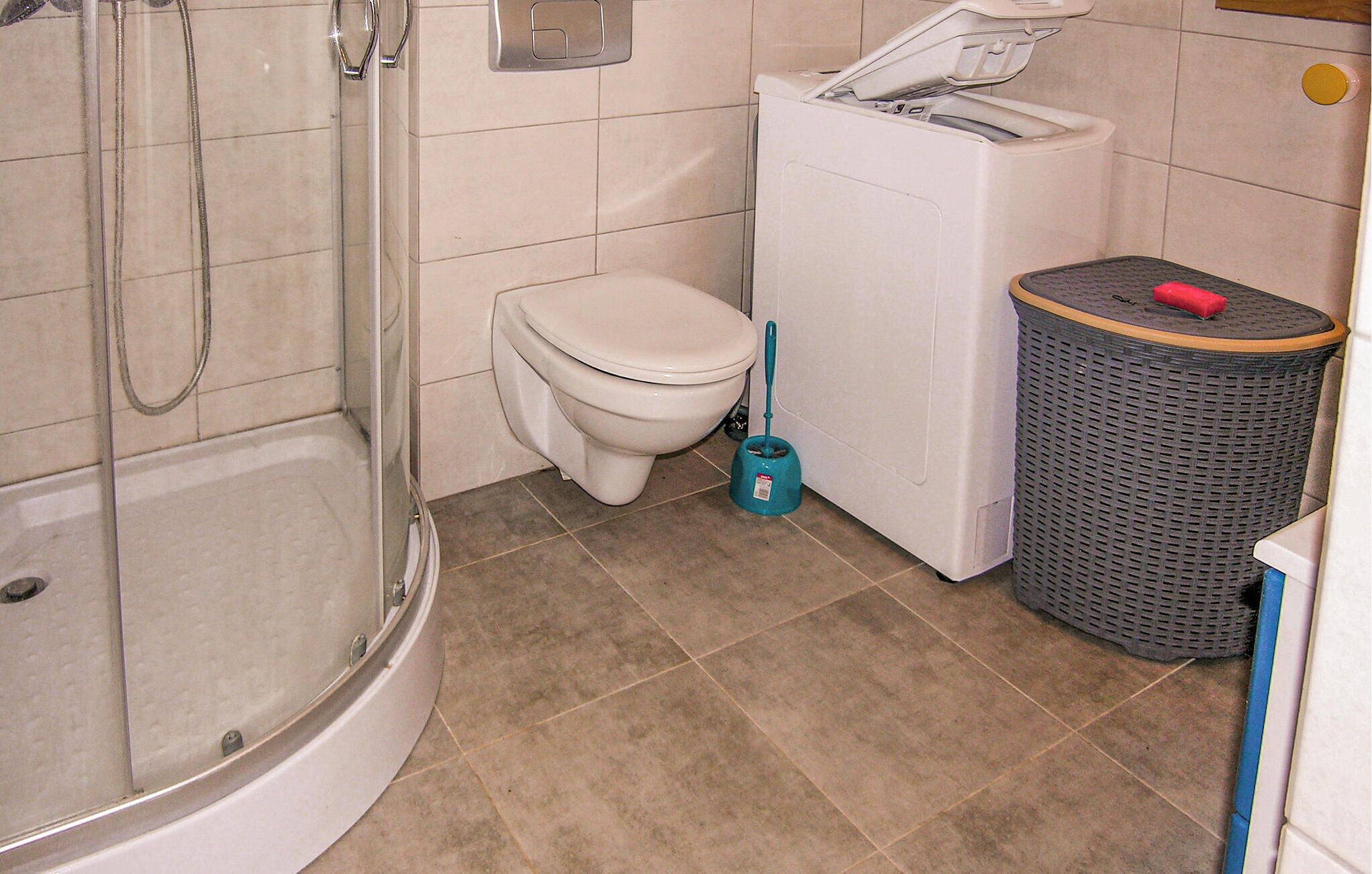 A bathroom with a shower, toilet, washing machine, and a trash bin.
