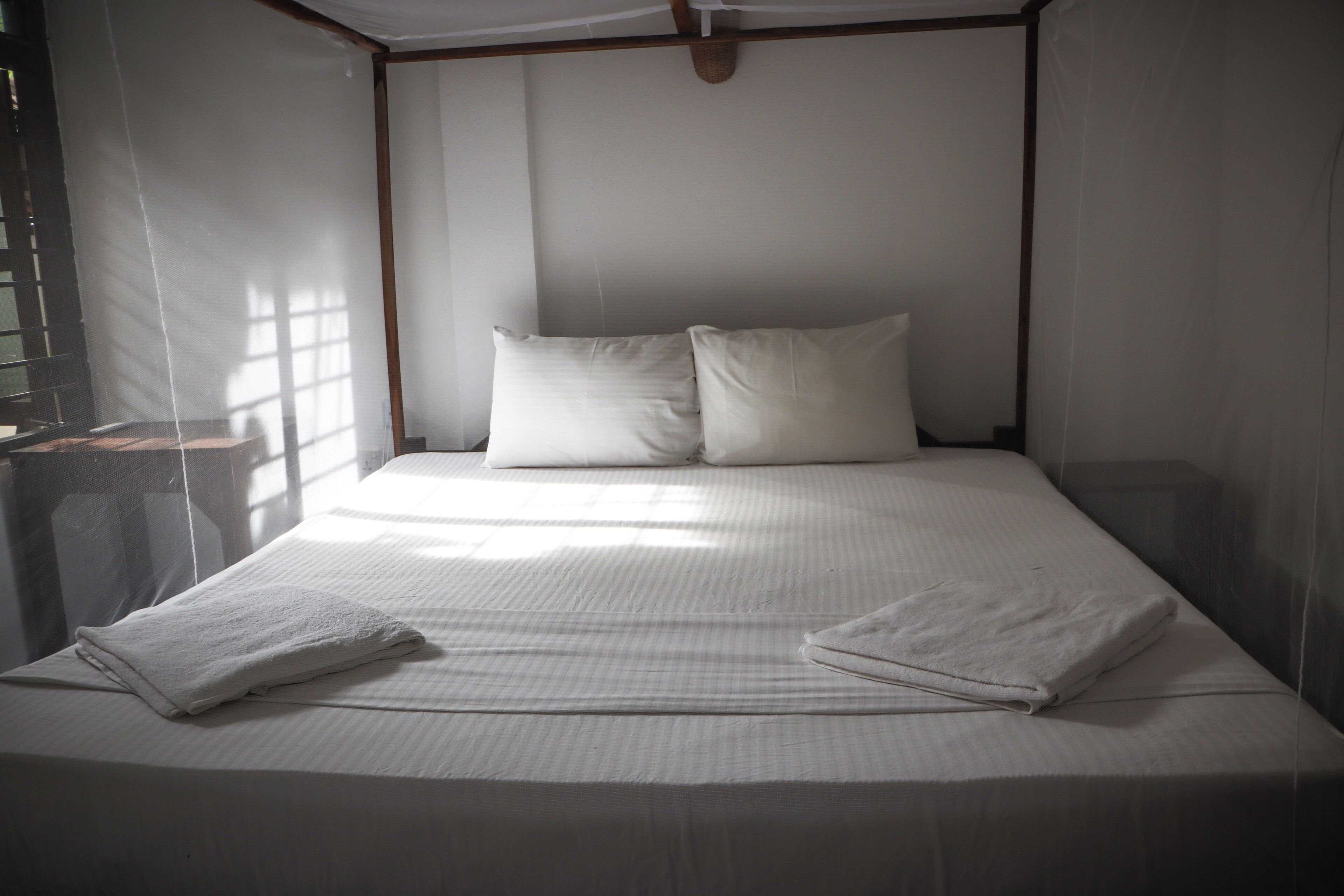 A bed with two pillows, a folded towel, and a window with natural light.