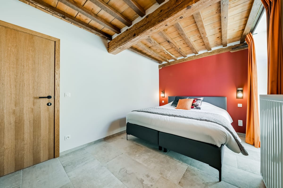 A bedroom with a wooden ceiling, a bed with white bedding, a red accent wall, and a window with orange curtains.
