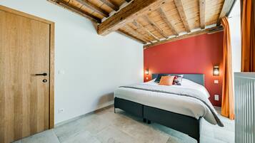 A bedroom with a wooden ceiling, a bed with white bedding, a red accent wall, and a window with orange curtains.