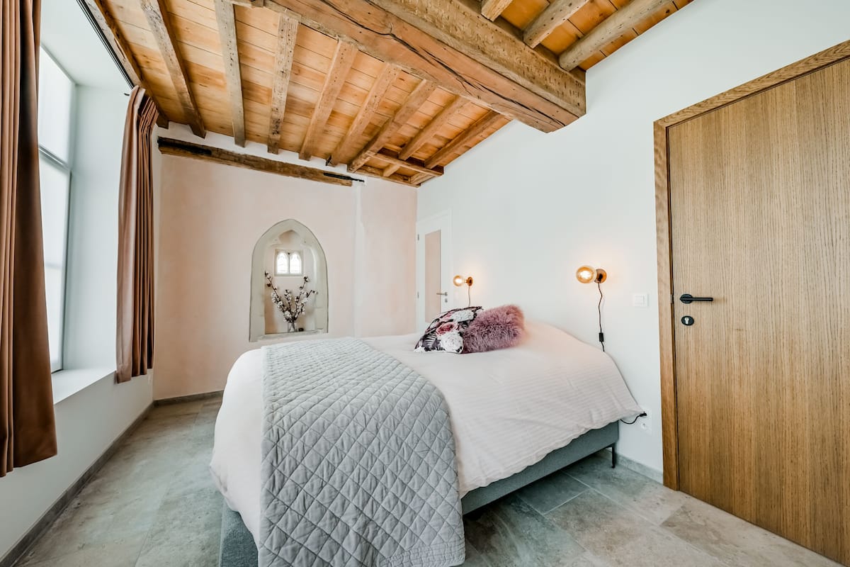 A bedroom with a bed, wooden ceiling beams, a door, and a window with curtains.