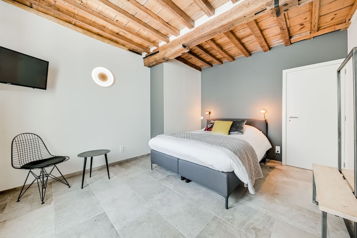 A bedroom with a bed, a chair, a small table, a TV, and a ceiling with exposed beams.