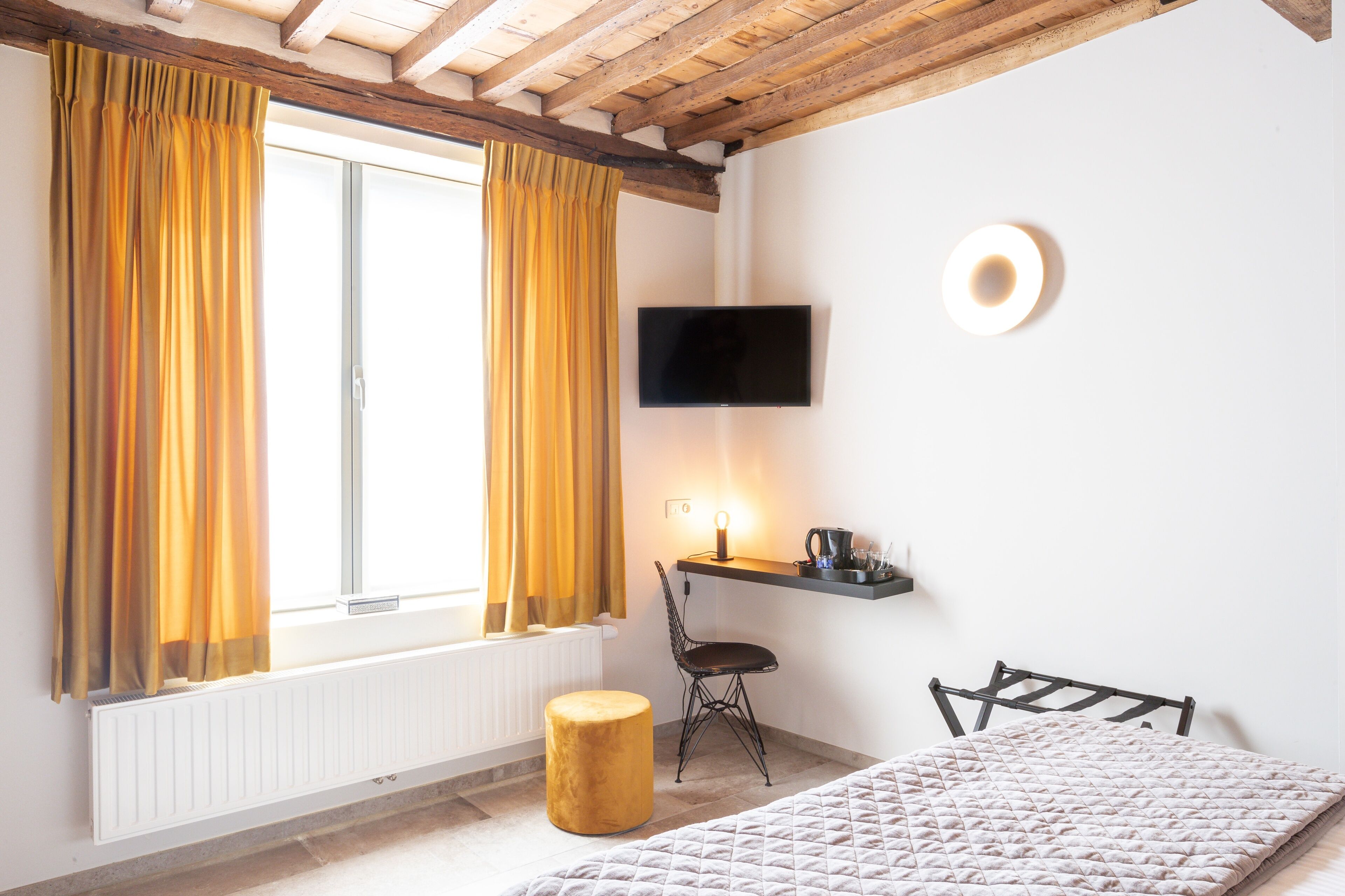 Une chambre à coucher avec un lit, une chaise, une petite table avec une lampe, un téléviseur et une fenêtre avec des rideaux.