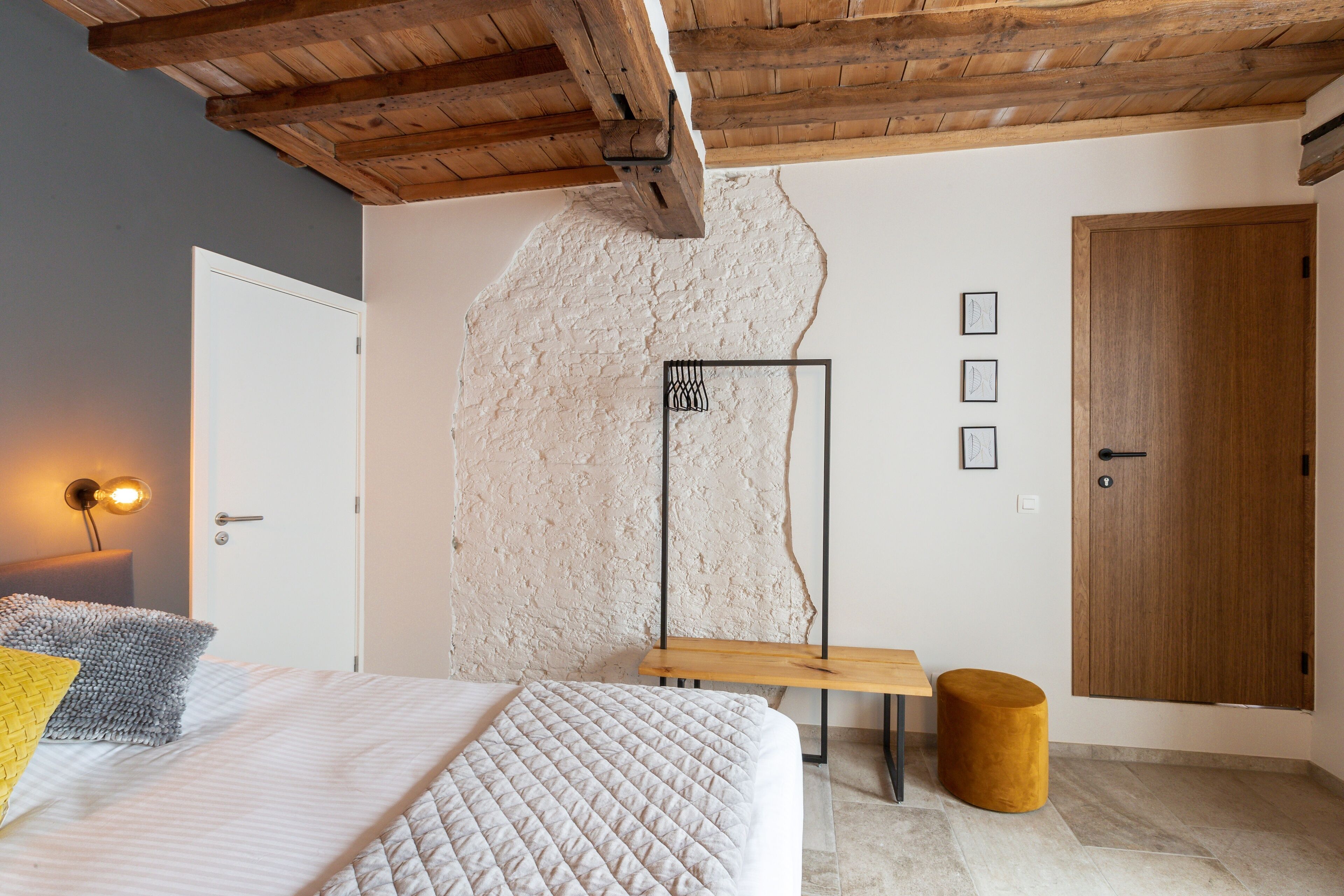 Een slaapkamer met een bed, een houten bankje, een rond voetenbankje, een deur en een gestructureerde muur.