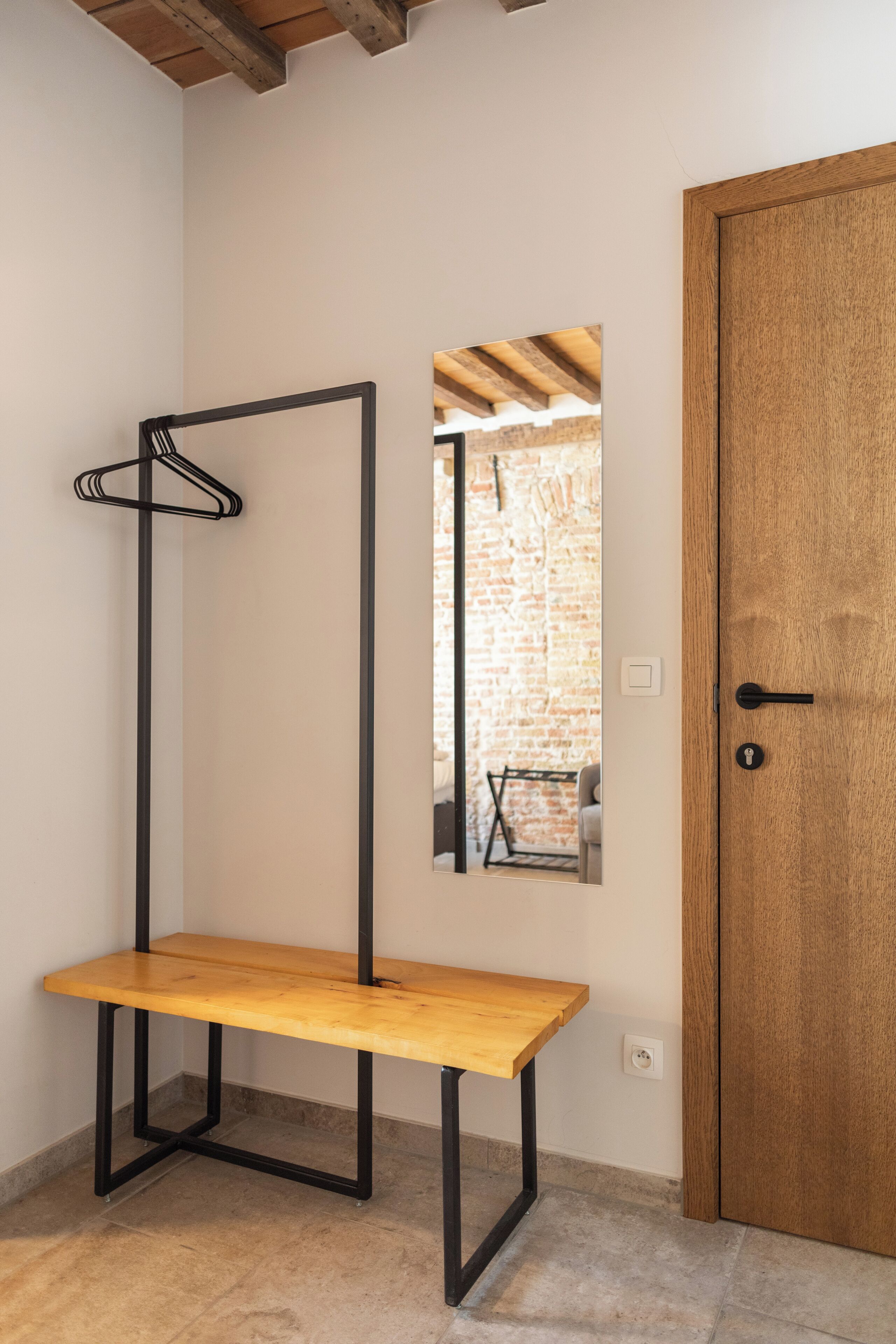 Quarto com um banco de madeira, um cabideiro, uma porta de madeira e um espelho refletindo uma parede de tijolos e uma mesa.