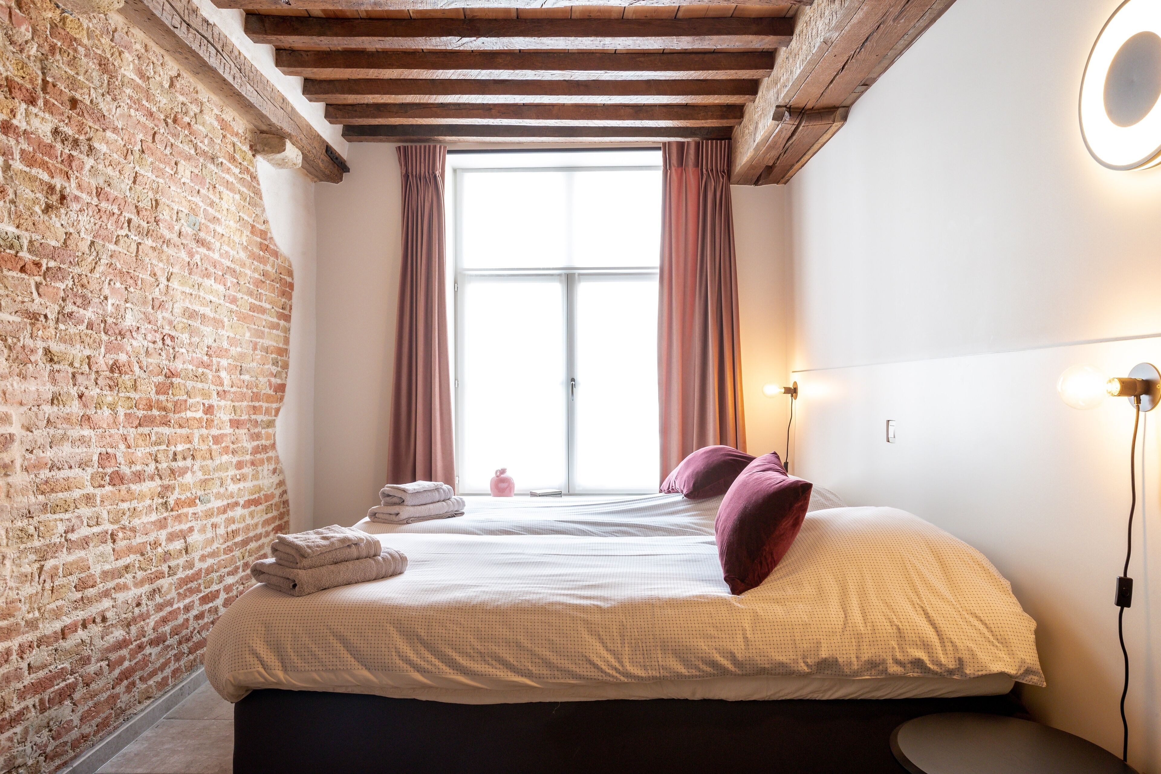Un dormitorio con pared de ladrillo, una ventana grande con cortinas, una cama con almohadas y una mesita de noche con lámpara.