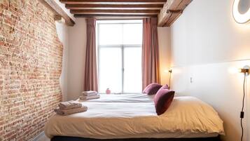 Un dormitorio con pared de ladrillo, una ventana grande con cortinas, una cama con almohadas y una mesita de noche con lámpara.