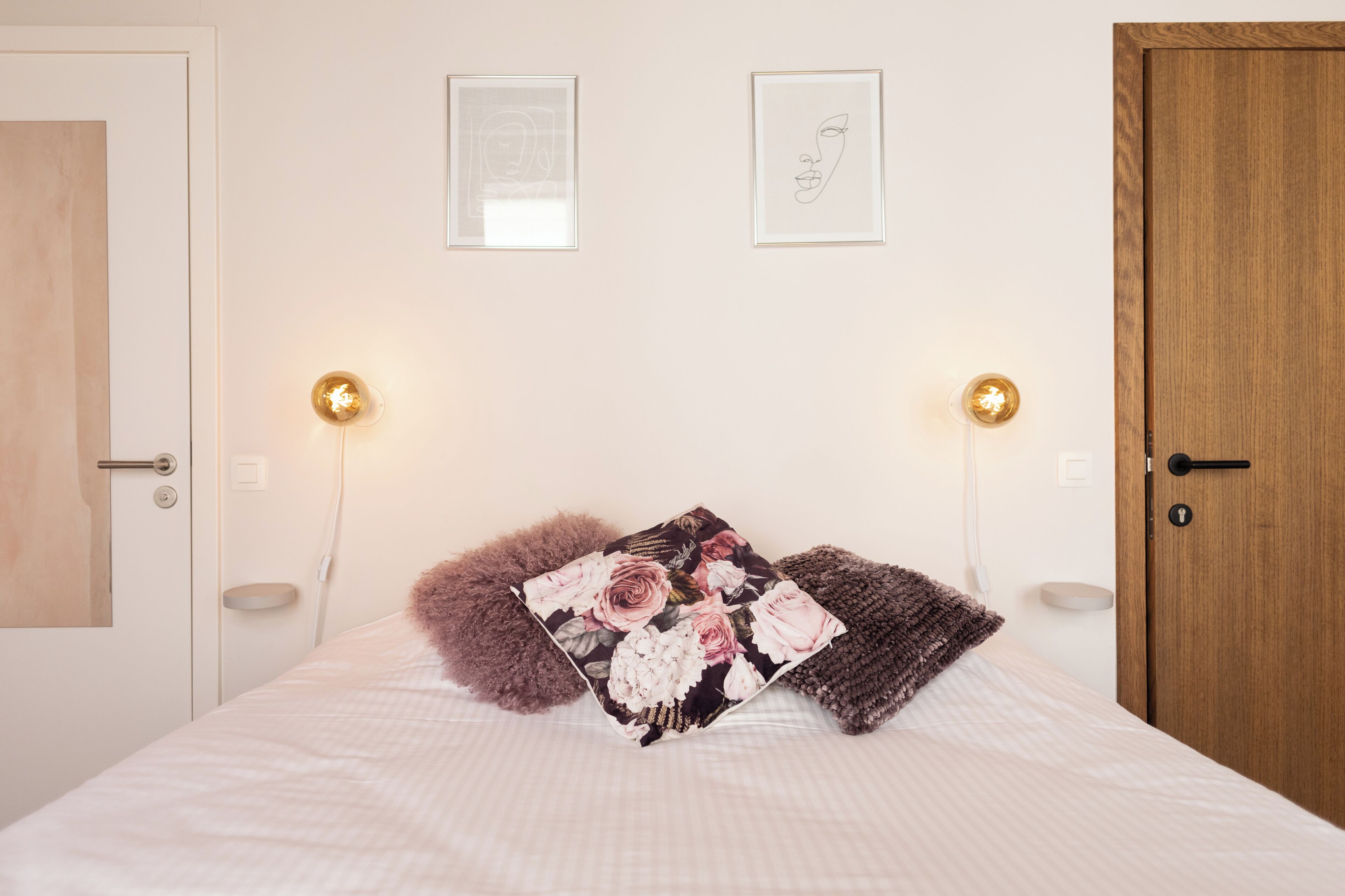 Een opgemaakt bed met bloemenkussens, twee ingelijste kunstwerken aan de muur en bedlampjes.