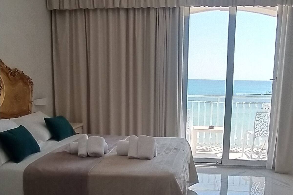 Habitación de hotel con una cama grande, un balcón con vista al mar y suelo de mármol.