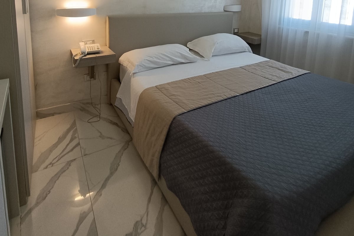 Habitación de hotel con cama, mesita de noche con lámpara, teléfono y ventana con cortinas.