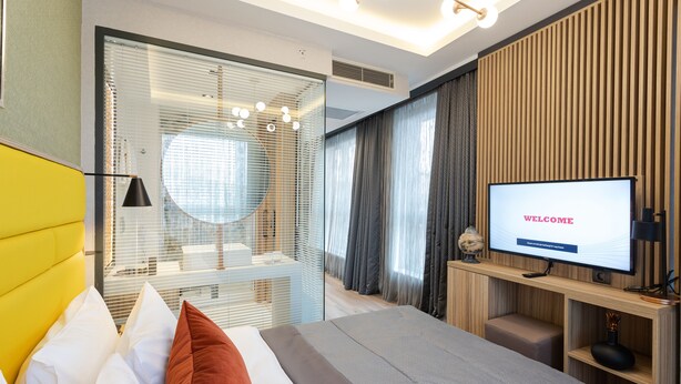 A modern hotel room with a large bed, a television displaying "WELCOME", and a bathroom with a glass partition. - ROX Hotel Ankara (Ankara)