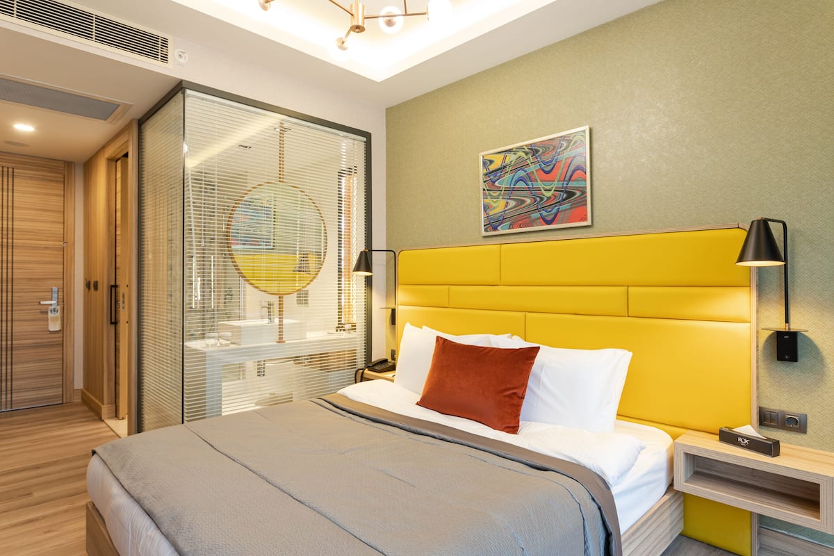 Una habitación de hotel moderna con una cama grande, un separador de vidrio y un cuadro abstracto llamativo en la pared.