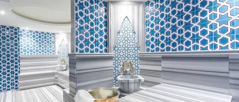 Un baño moderno con patrones geométricos de azulejos azules, suelo de mármol y una fuente decorativa central.