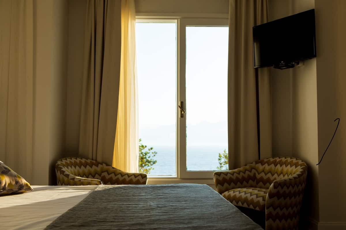 Habitación de hotel con cama, dos sillones, televisor y un ventanal con vista al mar.