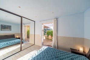 A bedroom with a bed, a bedside table with a lamp, a large mirror, and a sliding door leading to an outdoor area.