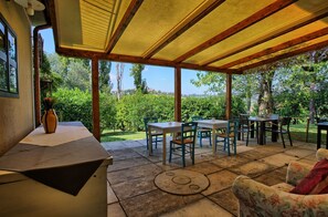 Un patio couvert avec des tables et des chaises, une horloge posée sur le sol et une vue sur la verdure et les arbres.