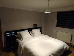 A bedroom with a bed, a bedside table, a hanging light fixture, a window with blinds, and a radiator.