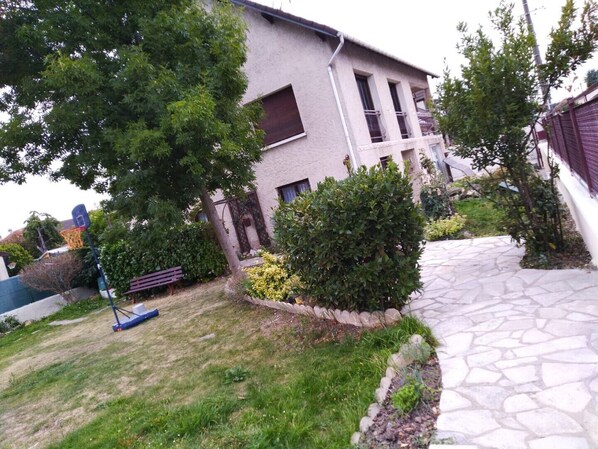 A residential building with a paved driveway, a basketball hoop, and a well-maintained garden with various shrubs and a bench.