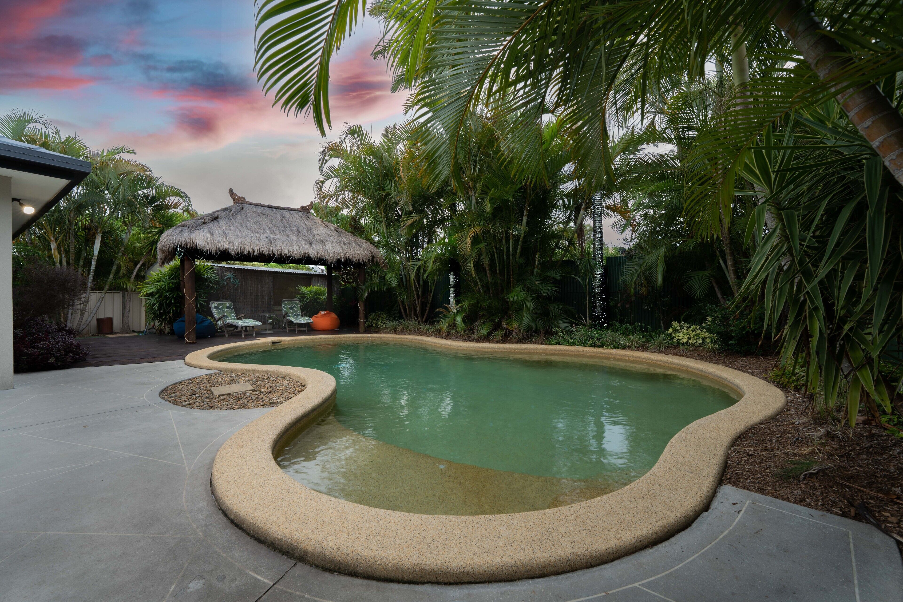 A swimming pool with a curved edge, surrounded by palm trees and a thatched-roof structure.
