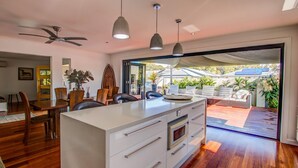 A modern kitchen with a central island, a dining area with wooden furniture, and a view of a backyard with plants and a patio. - Pets Allowed Burleigh Beach Pad (Burleigh Waters)
