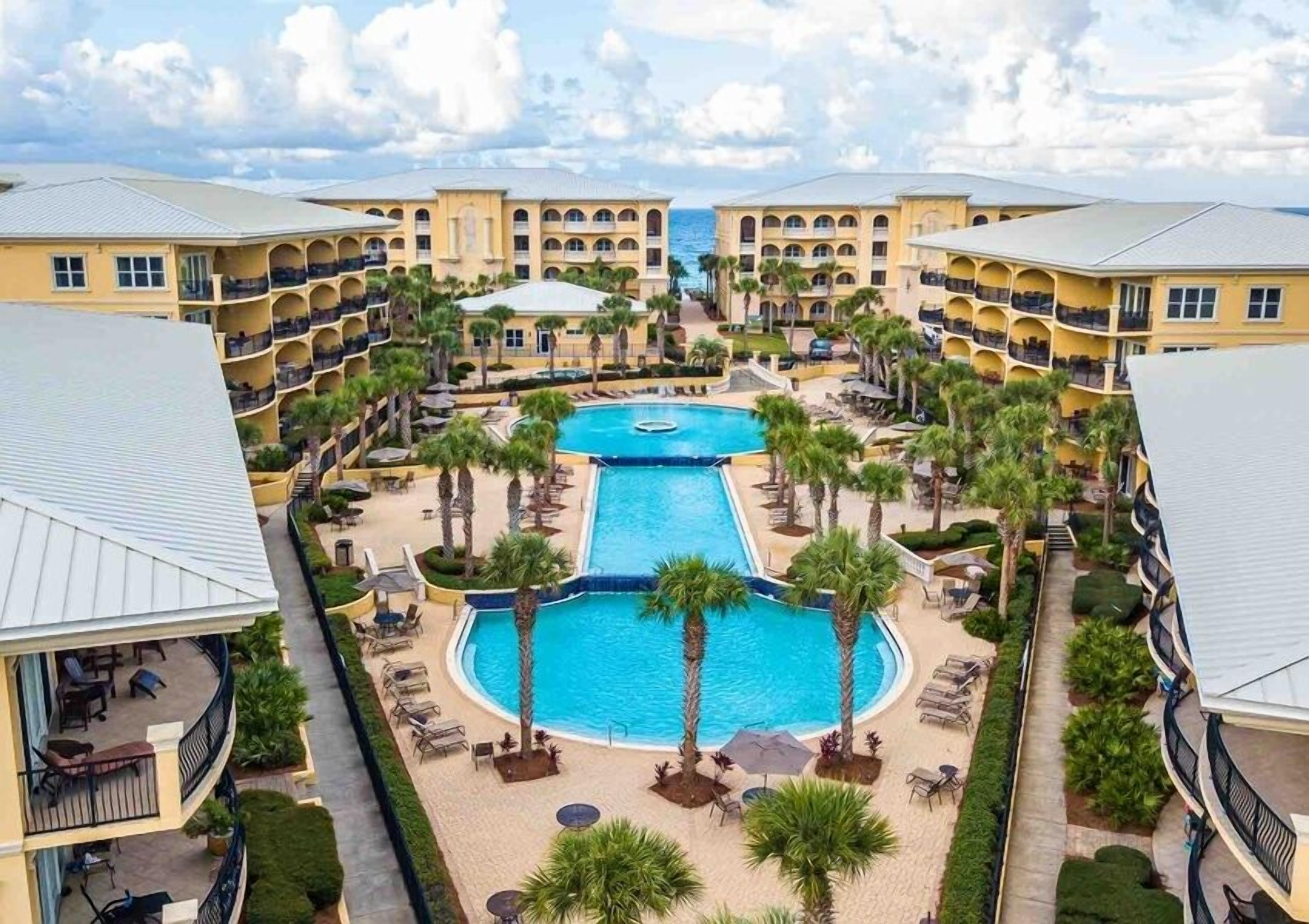 A resort with a central swimming pool surrounded by palm trees and lounge chairs, connected to multi-story buildings with balconies.