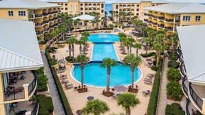 A resort with a central swimming pool surrounded by palm trees and lounge chairs, connected to multi-story buildings with balconies. - Adagio F-202 - Must See Poolside Unit!! (Santa Rosa Beach)