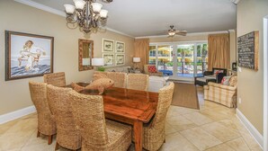 A spacious dining area with a wooden table and wicker chairs, a chandelier, and a view of the outdoor balcony. - Adagio F-202 - Must See Poolside Unit!! (Santa Rosa Beach)