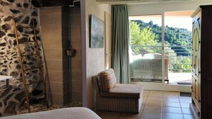 Un dormitorio con pared de piedra, una cama, una silla de mimbre y vistas a un balcón con una silla y una mesa.