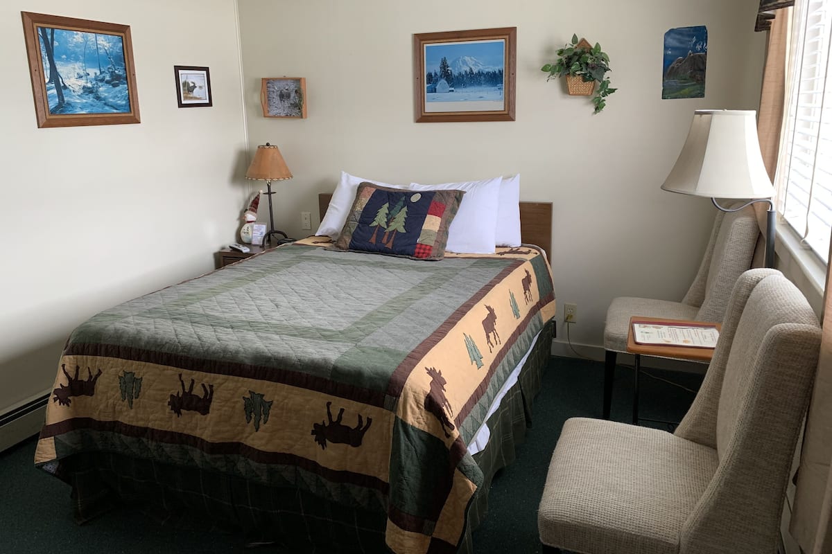 A hotel room with a bed, a chair, a nightstand, a lamp, and wall-mounted pictures.