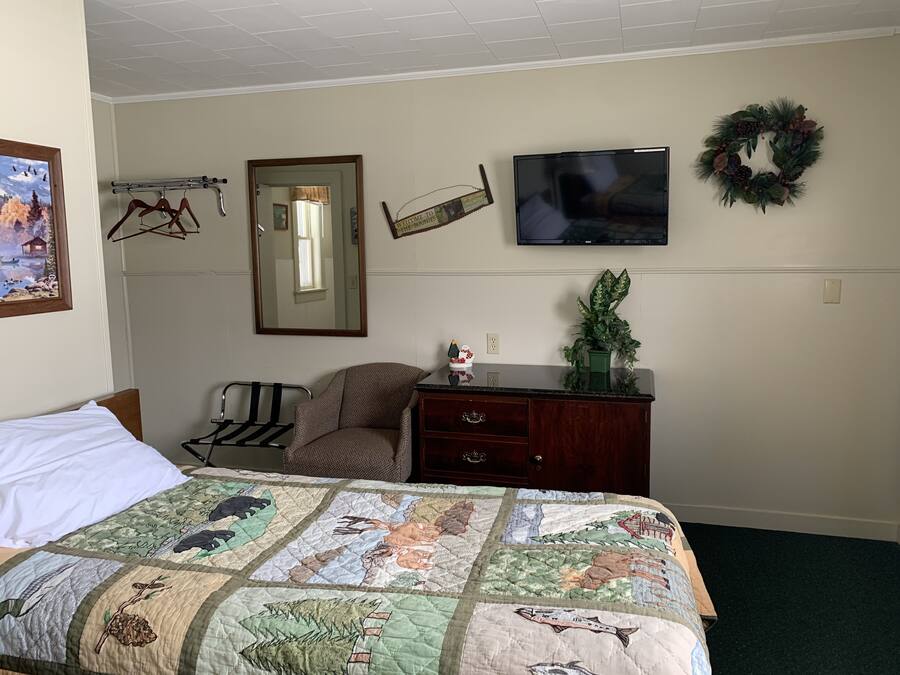A bedroom with a bed, a dresser, a chair, a TV, a mirror, and a wall with a wreath.