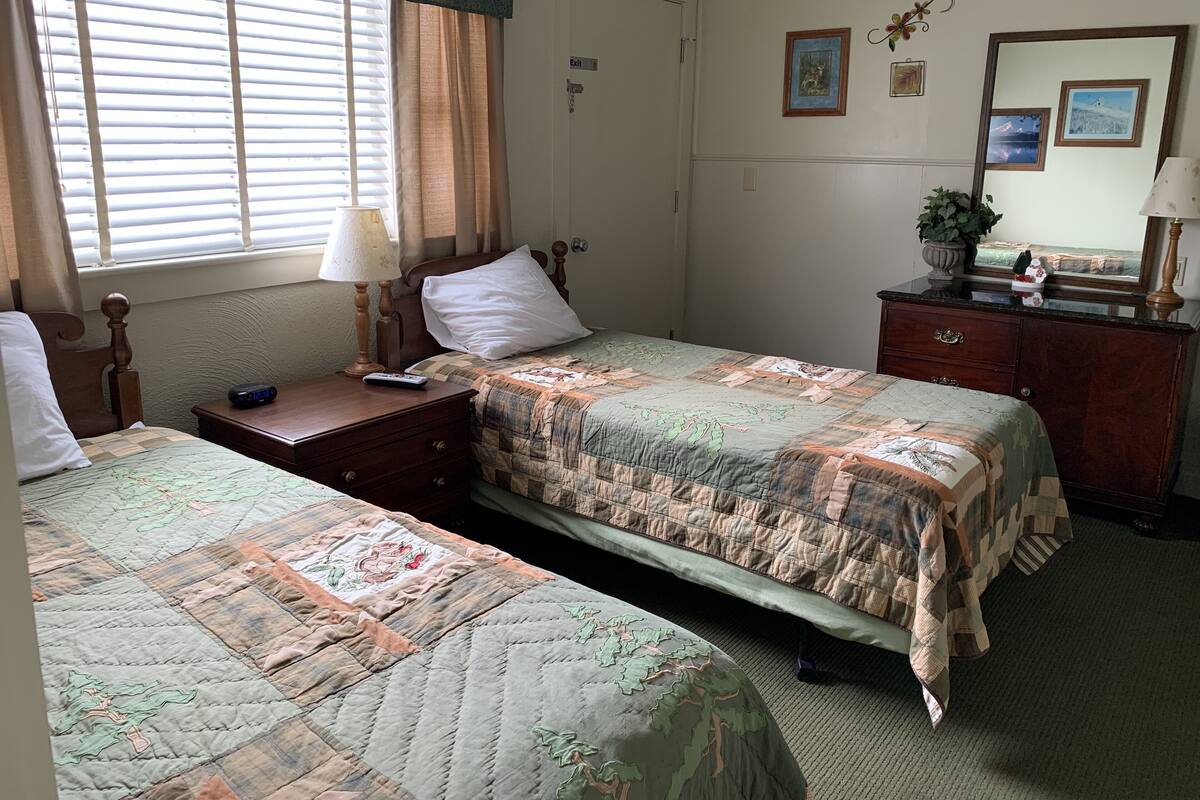A room with two single beds, a nightstand with a lamp, a dresser, and a window with blinds.