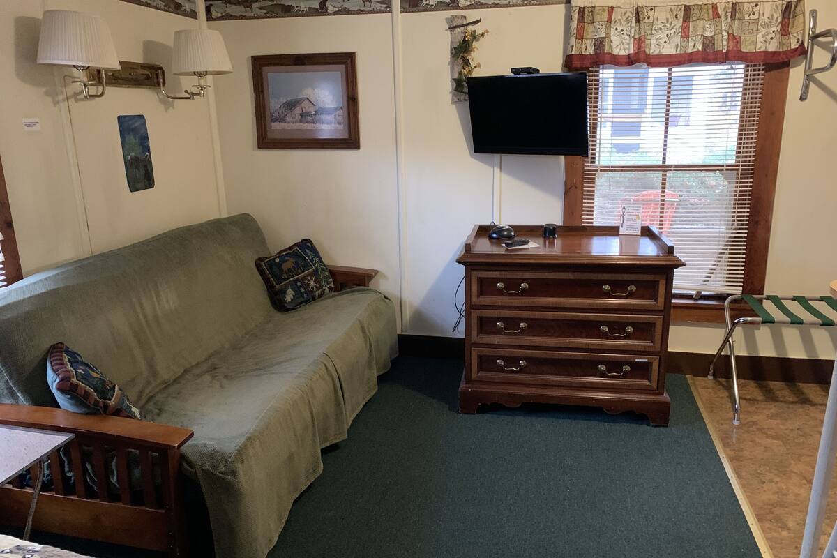 A room with a sofa, a wooden dresser, a television, and a window with blinds.
