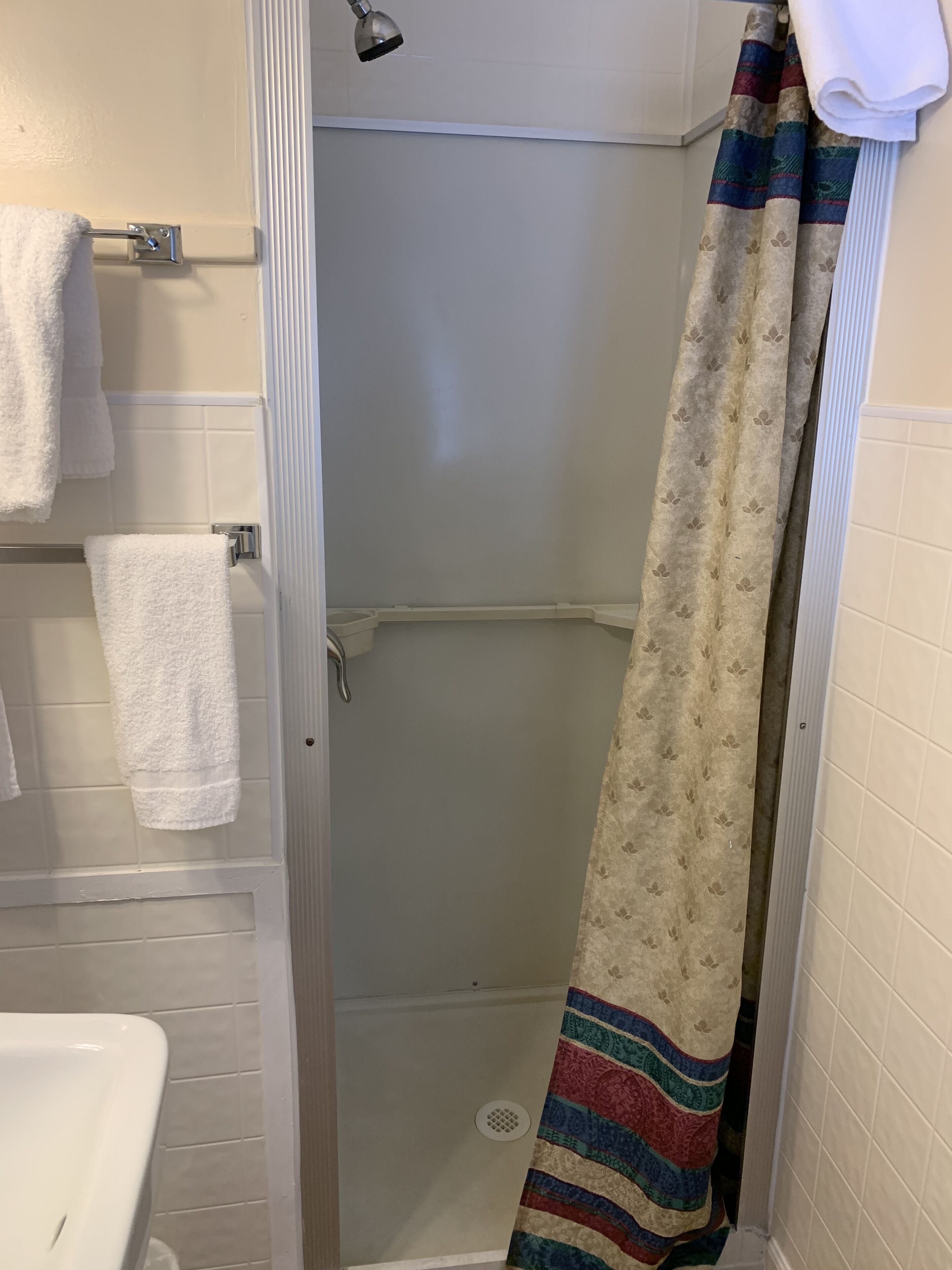 A bathroom with a shower, white tiled walls, a white sink, and a towel rack with white towels.