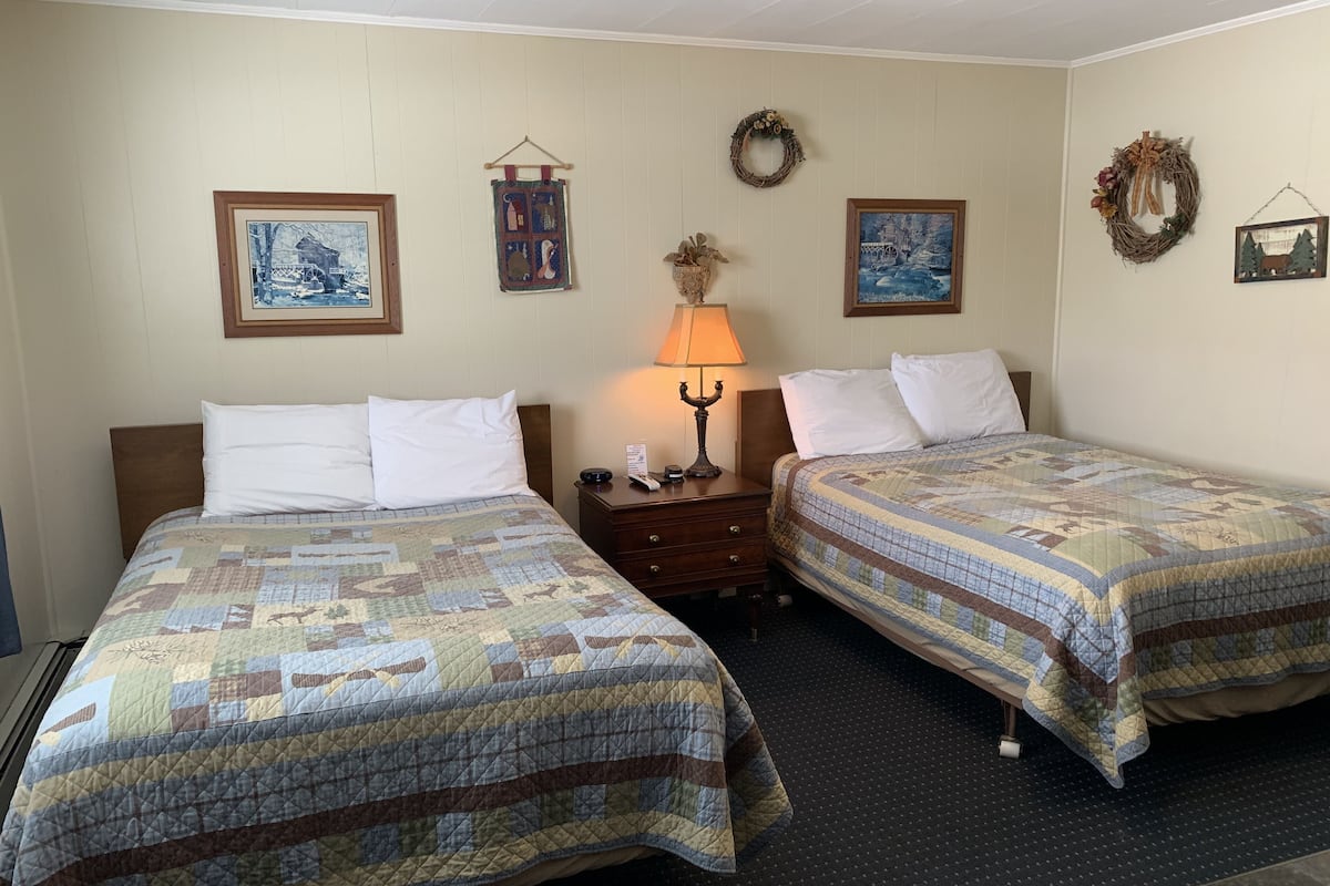 A hotel room with two beds, a nightstand, a lamp, and wall decorations including framed pictures and wreaths.