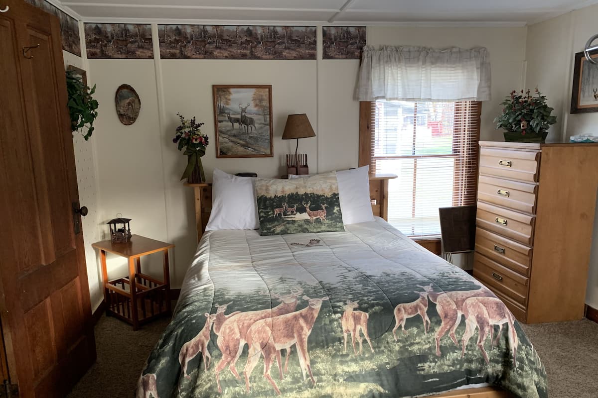 A bedroom with a bed featuring a deer print bedspread, a wooden dresser, a nightstand with a lamp, and a window with blinds.