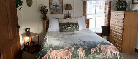 A bedroom with a bed featuring a deer print bedspread, a wooden dresser, a nightstand with a lamp, and a window with blinds.