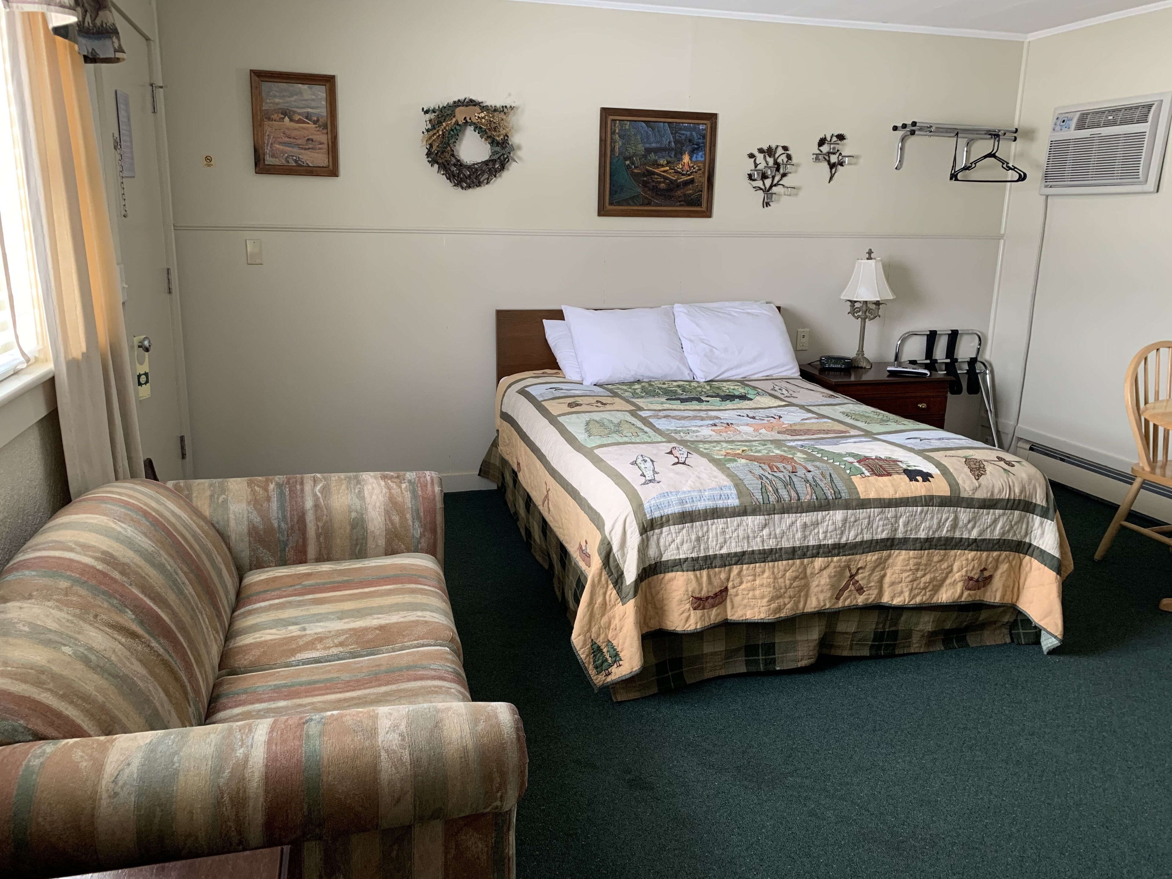 A bedroom with a bed, a chair, a nightstand, and a wall with a wreath and two framed pictures.
