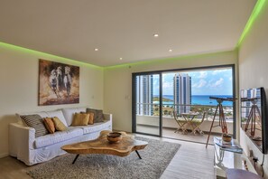 Ein modernes Wohnzimmer mit einem weißen Sofa, einem Holztisch und einem großen Gemälde. Durch die Glastüren ist ein Balkon mit Blick auf das Meer und Gebäude zu sehen.