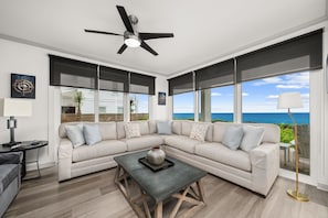 A modern living room with a sectional sofa, a coffee table, a ceiling fan, and large windows with a view of the ocean.