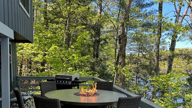 Eine Holzterrasse mit rundem Tisch und Stühlen, einer Feuerstelle und Blick auf ein bewaldetes Gebiet.