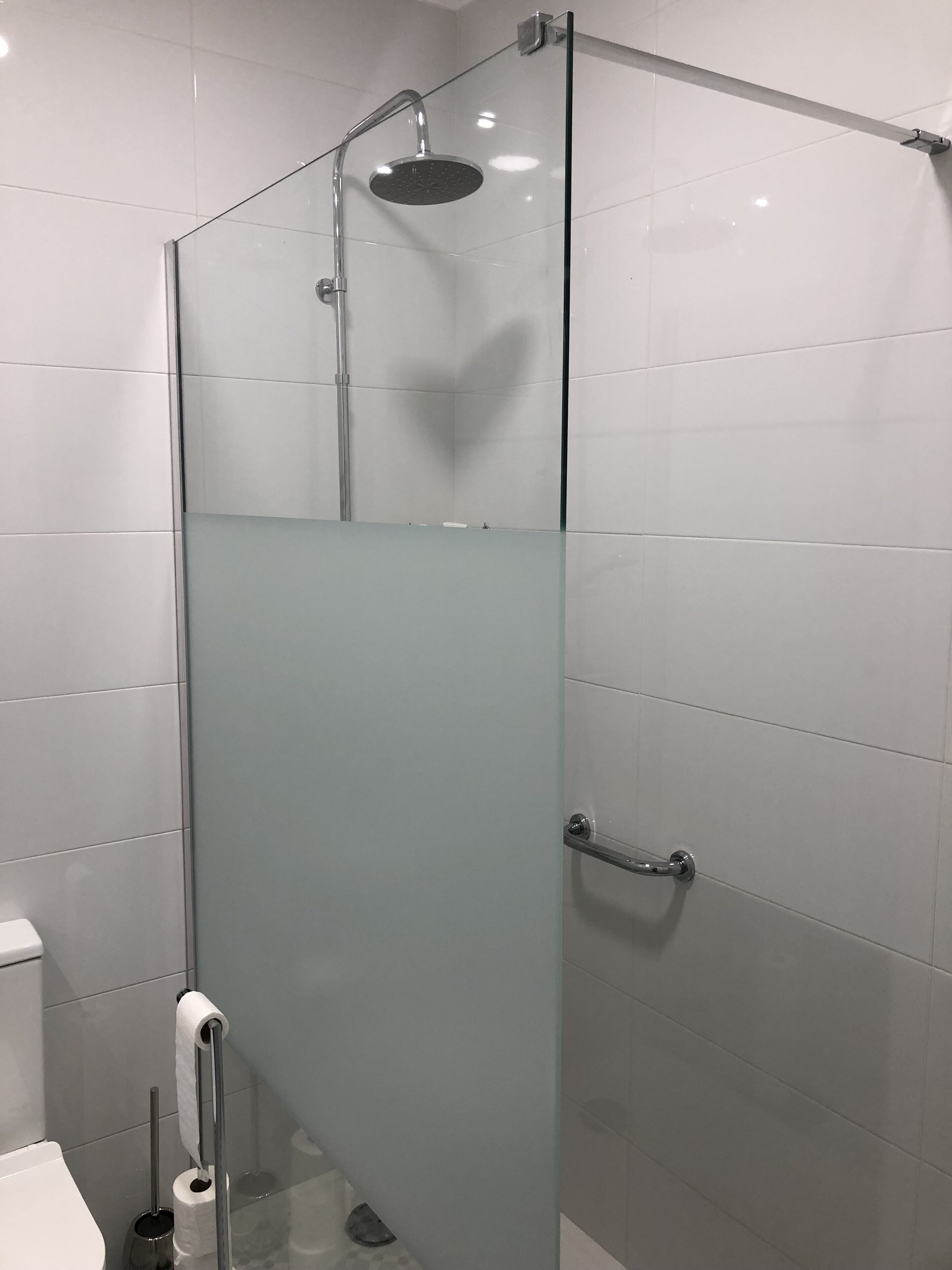 Une salle de bain moderne dotée d’une douche à parois de verre, de murs recouverts de carreaux blancs et de toilettes avec un porte-serviettes en papier.