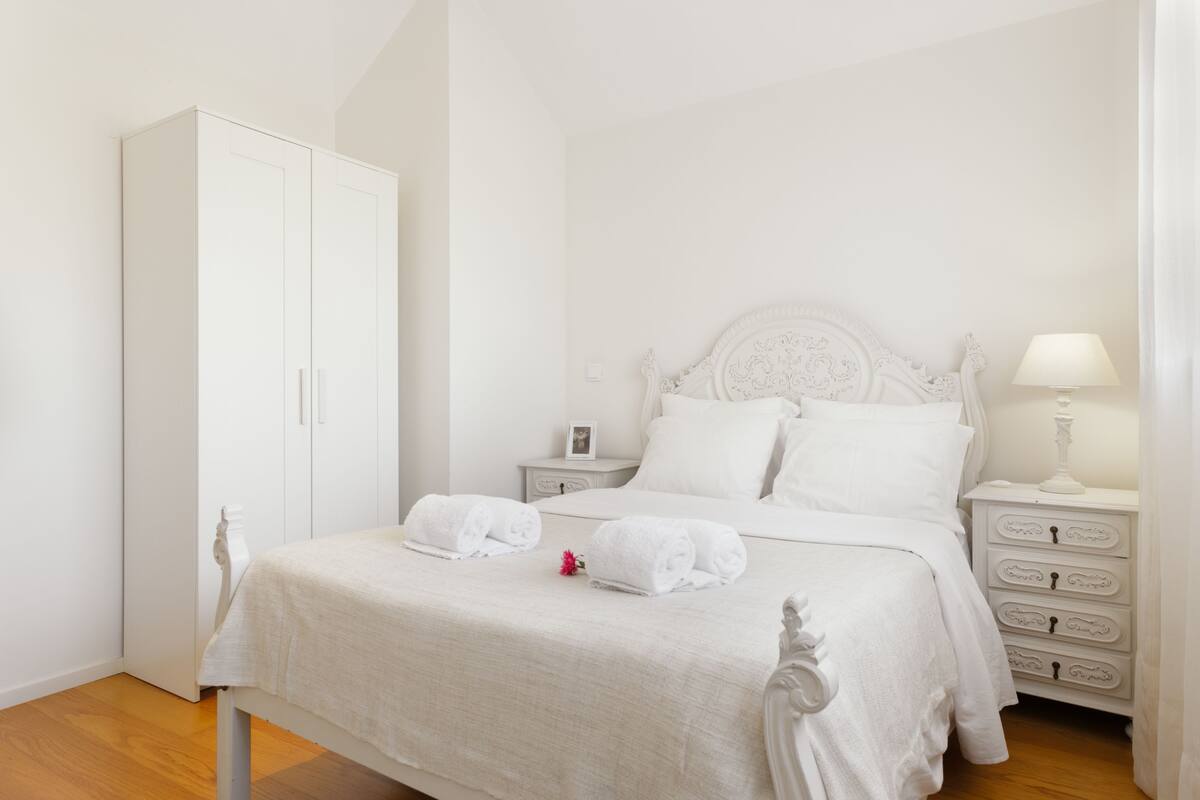 A neatly made bed with white linens and a decorative headboard, a white wardrobe, and a bedside table with a lamp.