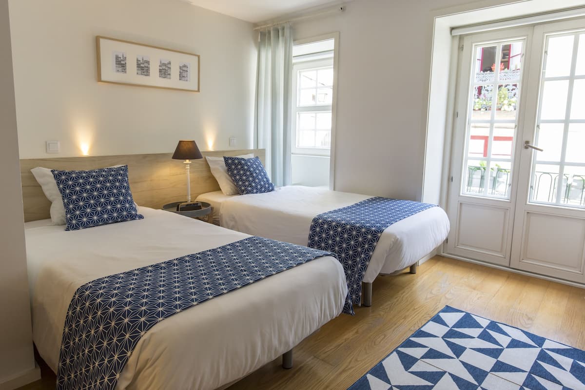 A hotel room with two beds, a nightstand, a lamp, a window with curtains, and a door leading to an outdoor area.