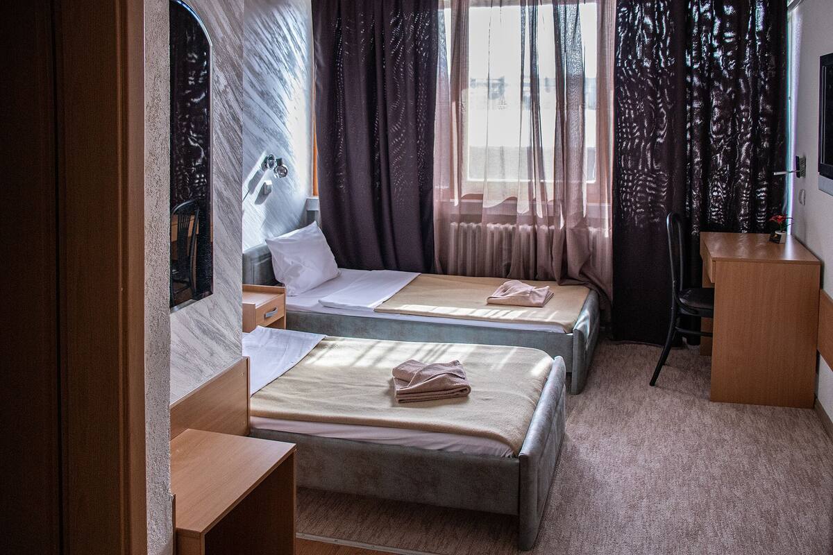 A compact hotel room with two single beds, a small desk, a chair, and a window with curtains.