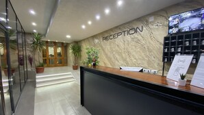 A reception area with a marble wall, a reception desk, potted plants, and a security monitor. - Hotel Moment (Velika Plana)