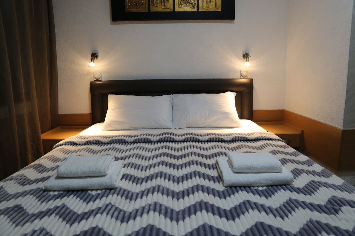 A neatly made bed with a patterned bedspread, two pillows, and two folded towels on a wooden nightstand.