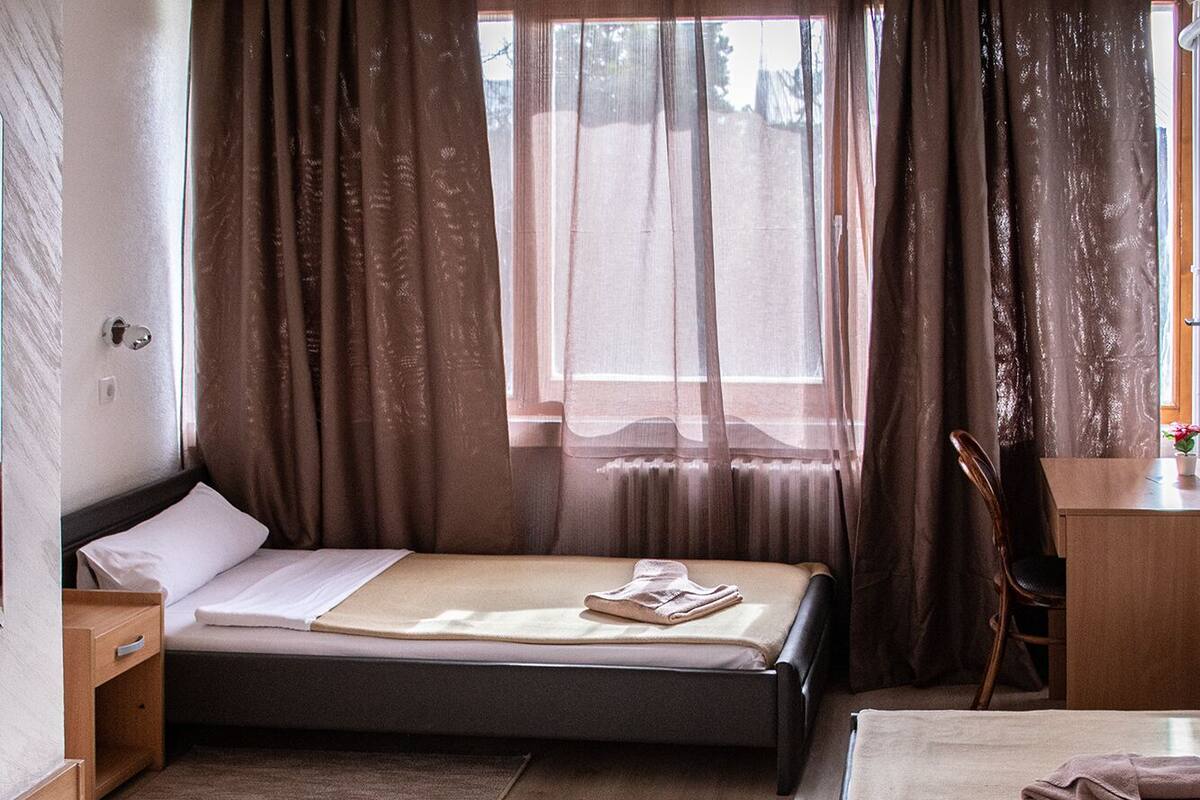 A compact hotel room with a single bed, a desk, a chair, a small storage unit, and a window with curtains.