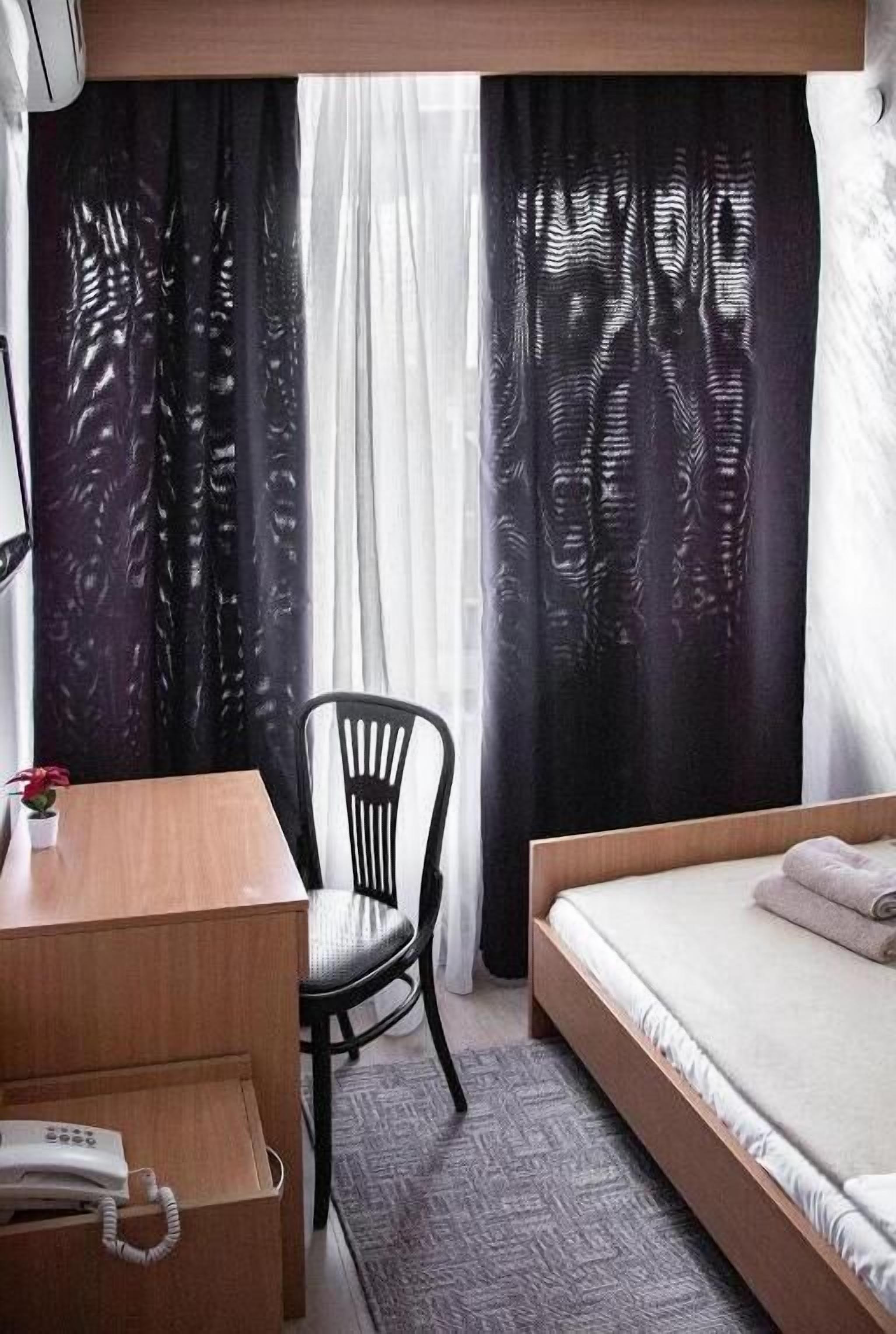 A hotel room with a bed, a desk with a telephone, a chair, and a window with patterned curtains.